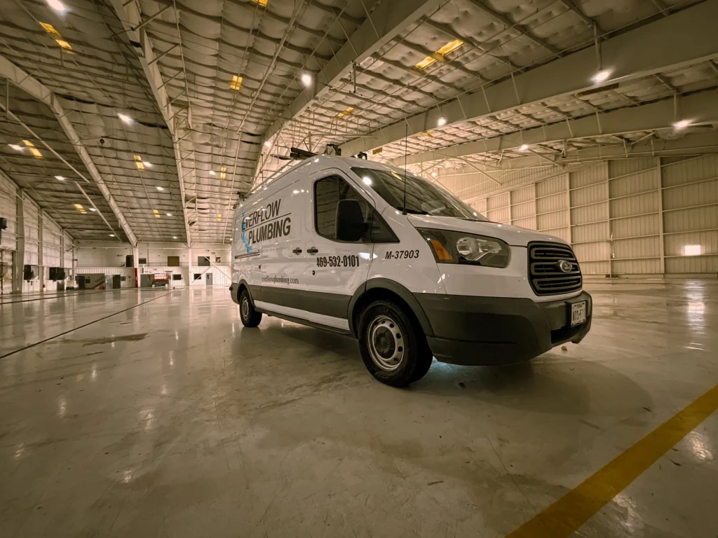 A white plumbing service van is parked inside a spacious, empty warehouse with high ceilings and bright overhead lights, ready to serve its broad service area.