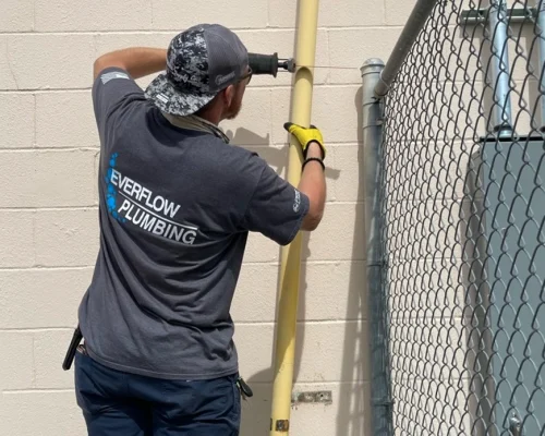 Everflow Technician Dallas – Fort Worth Texas A plumber wearing an "Everflow Plumbing" shirt works on a yellow pipe attached to a light-colored wall near a chain-link fence, showcasing the dedication you’ll read about us on our company page.